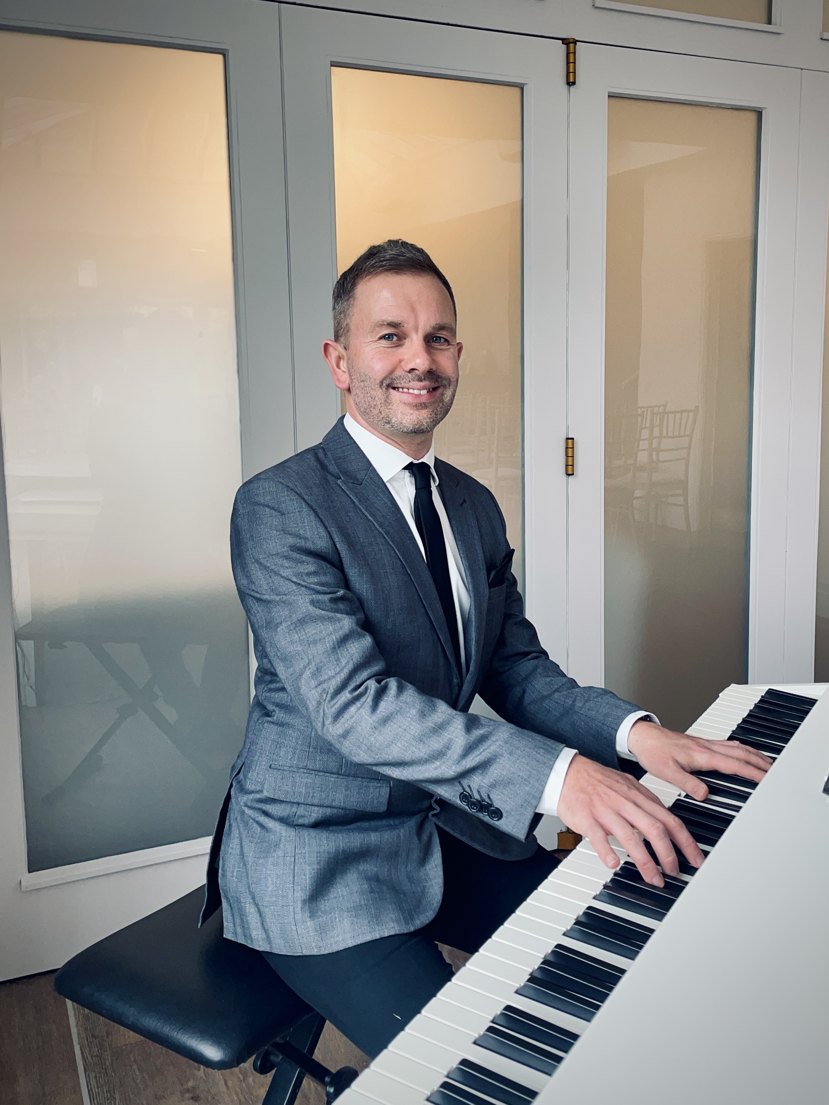 Wedding Pianist Craig Smith, AKA The Lake District Pianist, smiles at the camera while playing a white baby grand piano at a Broadoaks Country House wedding ceremony. He wears a charcoal jacket over a white shirt, black tie and black trousers.
