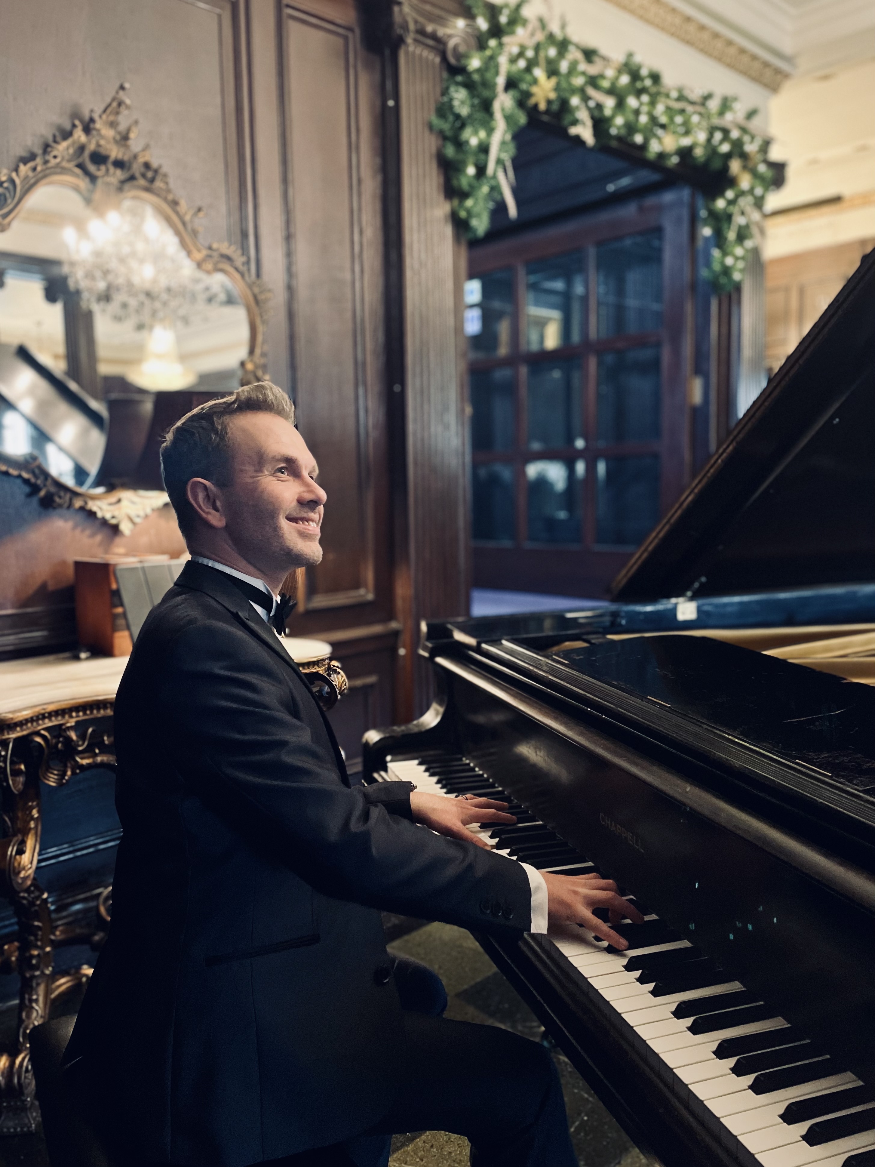 Craig Smith sits at the piano at Eaves Hall. He wears a black dinner jacket and bowtie and smiles at a wedding guest out of shot.