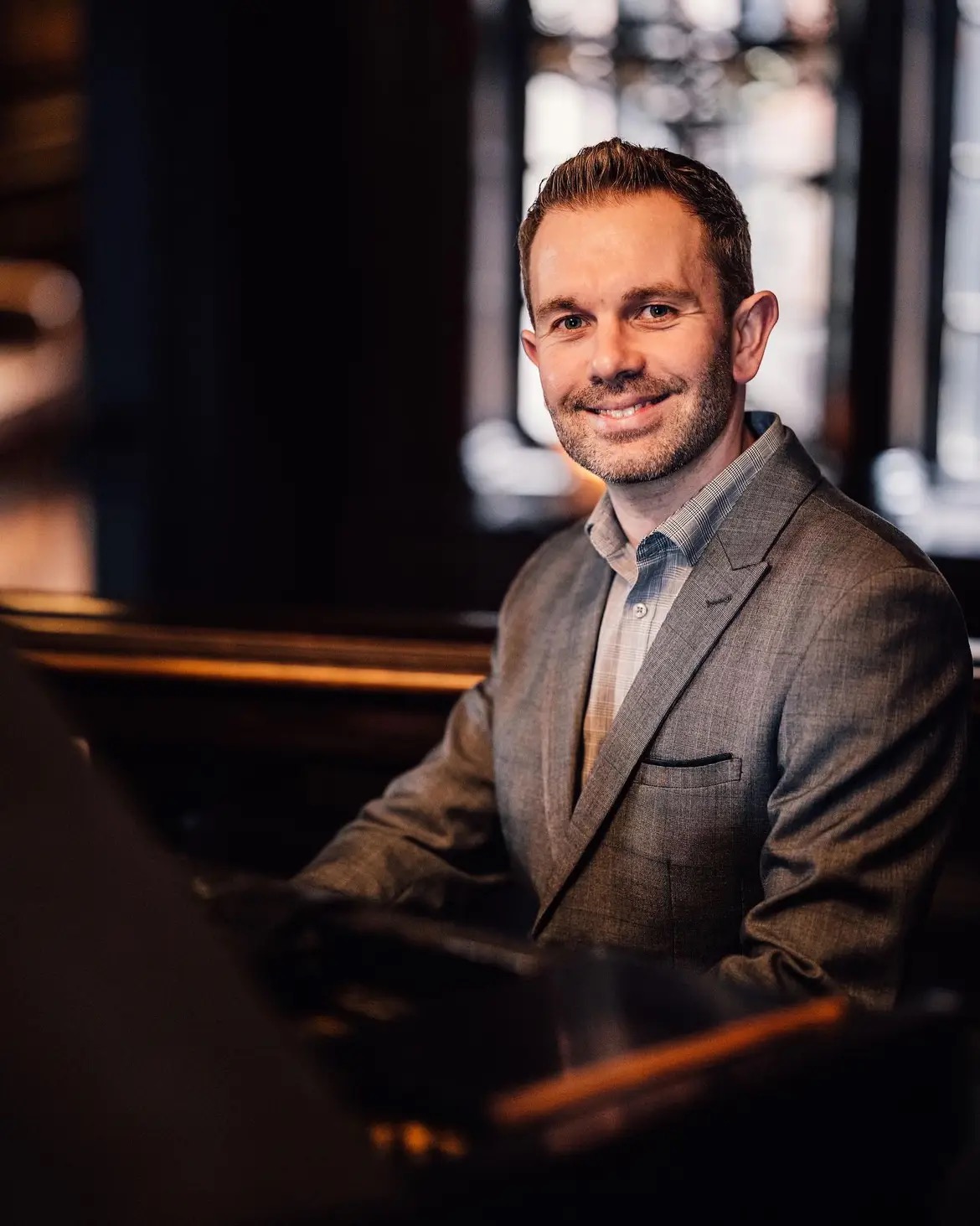 Wedding pianist Craig Smith smiles to the camera from behind the piano at an Eaves Hall wedding. He wears a grey jacket and light-coloured open-collared shirt