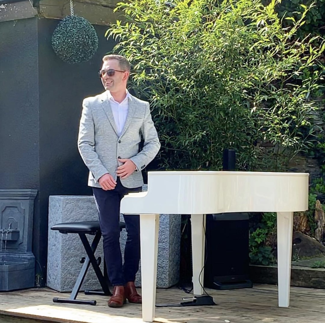 Wedding pianist Craig Smith smiles standing next to his white baby grand piano in the Sparth House gardens